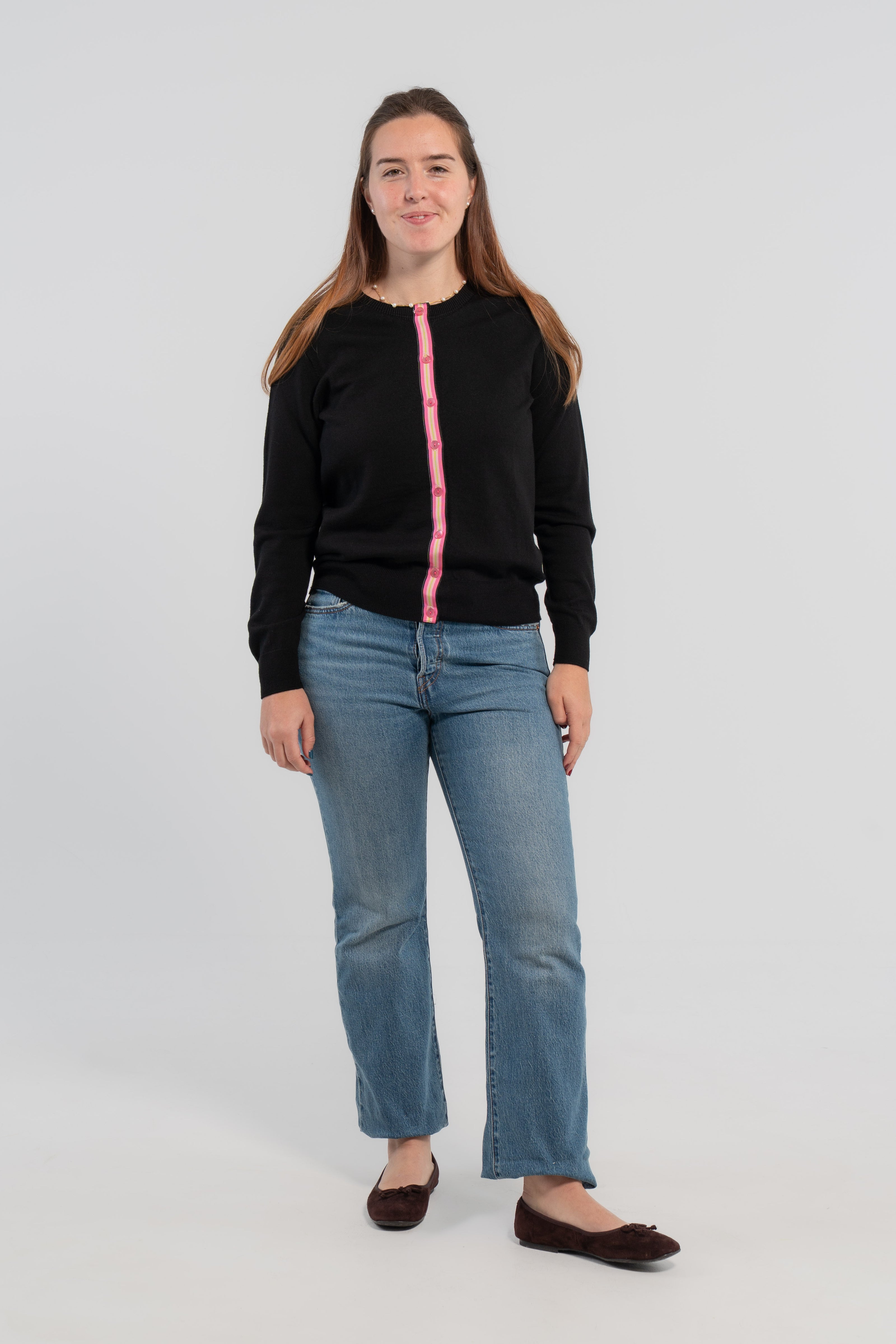 Woman wearing buttoned black cardigan with pink buttons and hot pink, light pink, and green striped grosgrain ribbon trim, jeans, ballet flats on plain background.