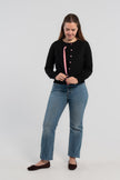 Woman wearing and buttoning black cardigan with pink buttons and hot pink, light pink, and green striped grosgrain ribbon trim over matching black sleeveless sweater, jeans, ballet flats and plain background.