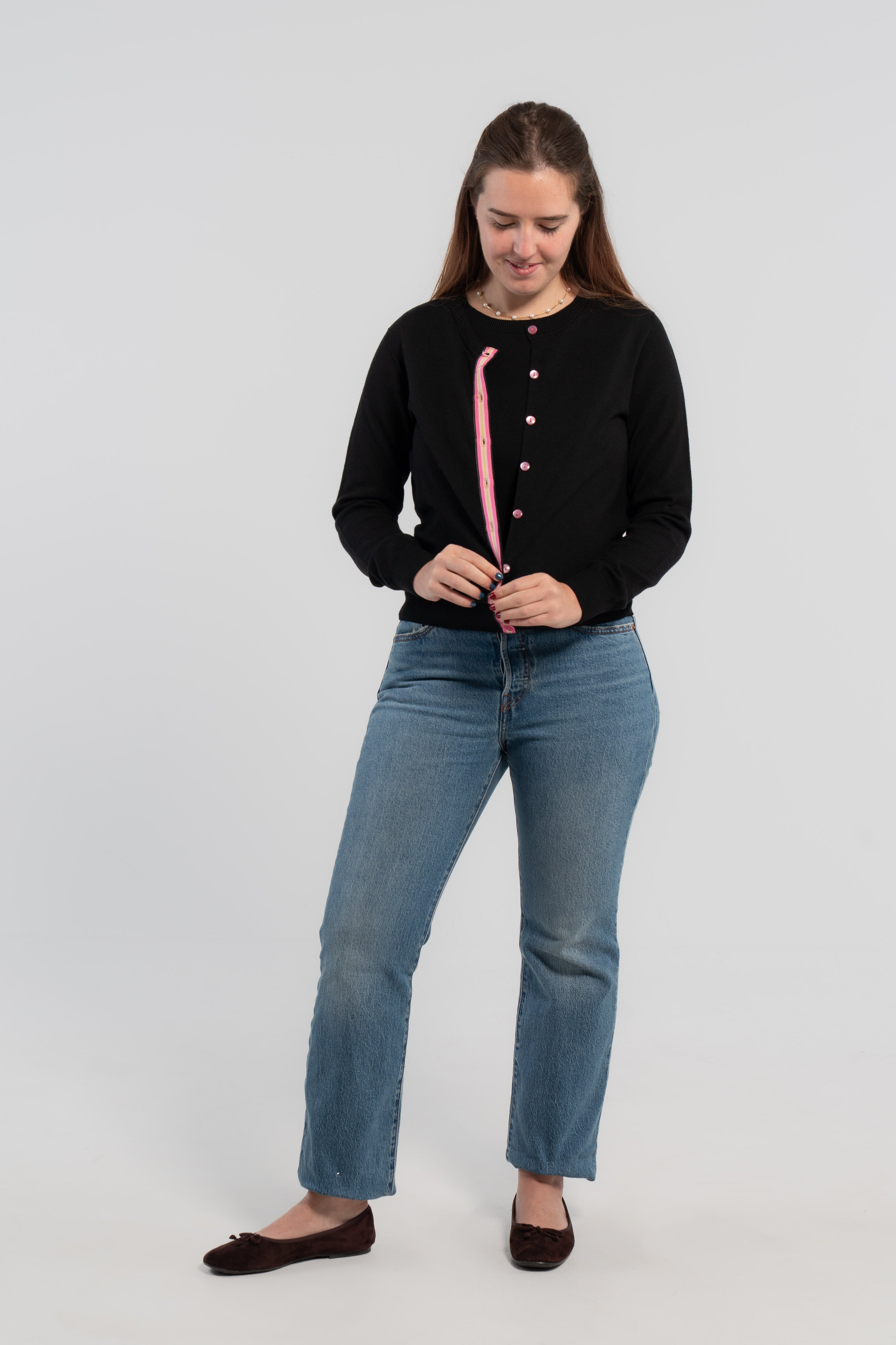 Woman wearing and buttoning black cardigan with pink buttons and hot pink, light pink, and green striped grosgrain ribbon trim over matching black sleeveless sweater, jeans, ballet flats and plain background.