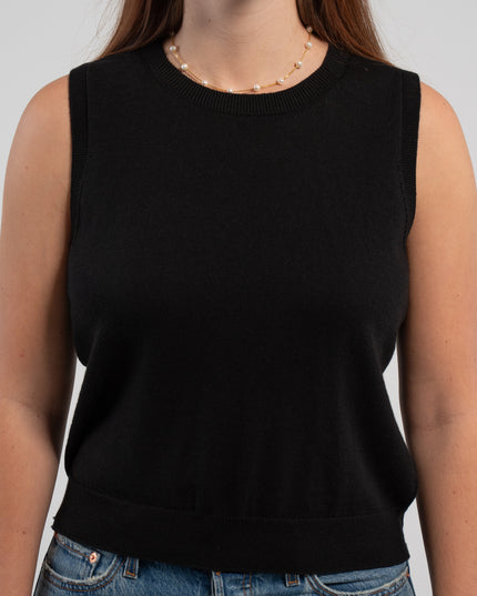Close up of woman wearing black sleeveless sweater, jeans, pearl choker neckless on plain background.