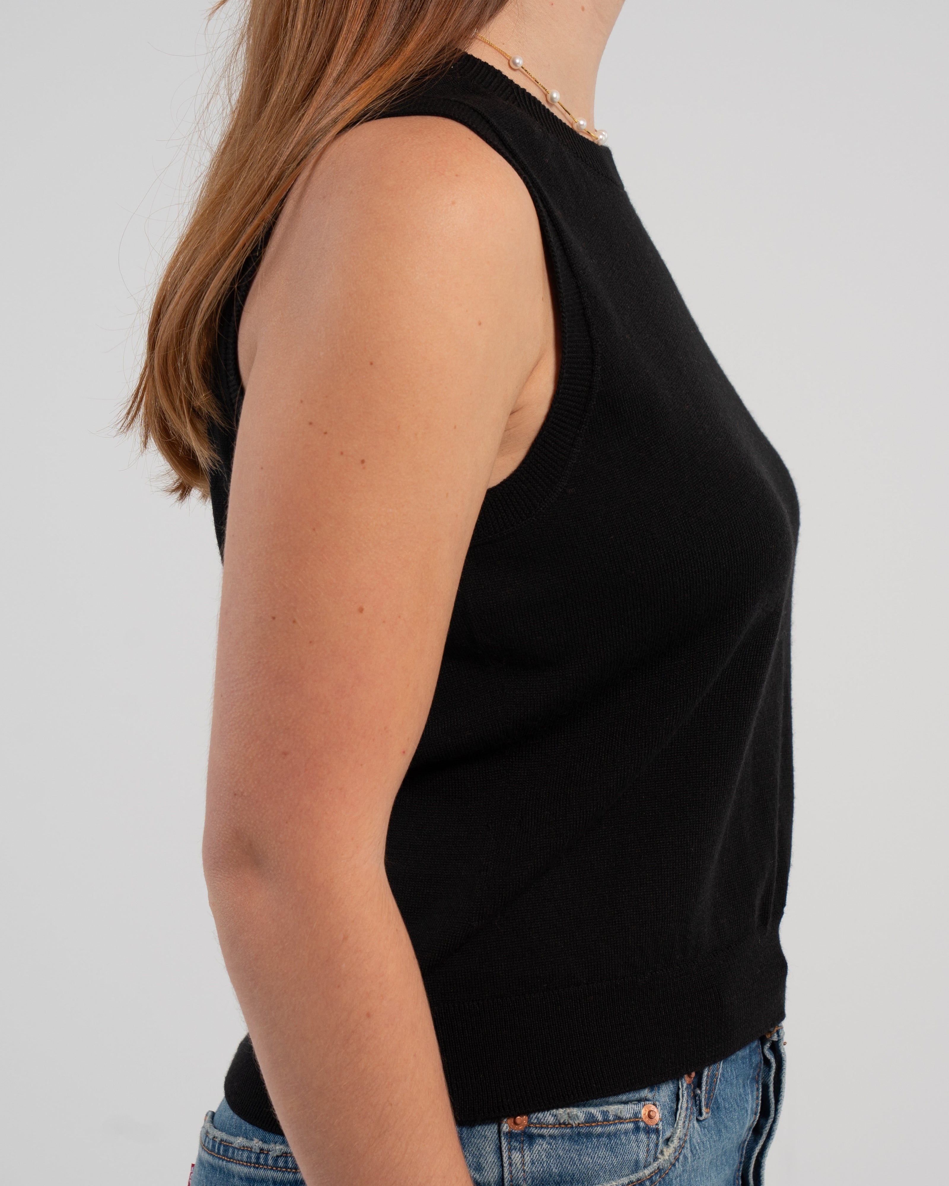 Sideview of woman wearing black sleeveless sweater, jeans, pearl choker neckless on plain background.