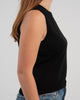 Sideview of woman wearing black sleeveless sweater, jeans, pearl choker neckless on plain background.