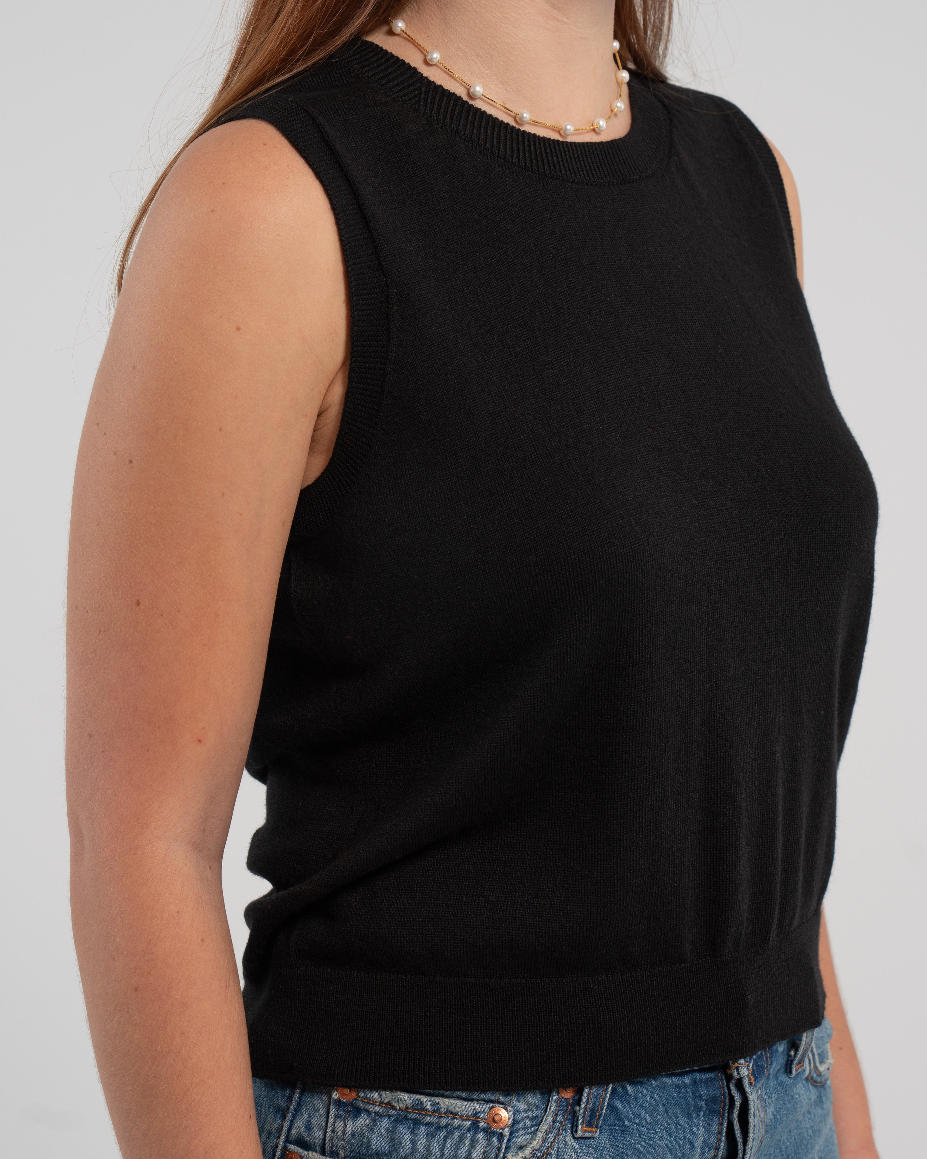 Close up of woman wearing black sleeveless sweater, jeans, pearl choker neckless on plain background.