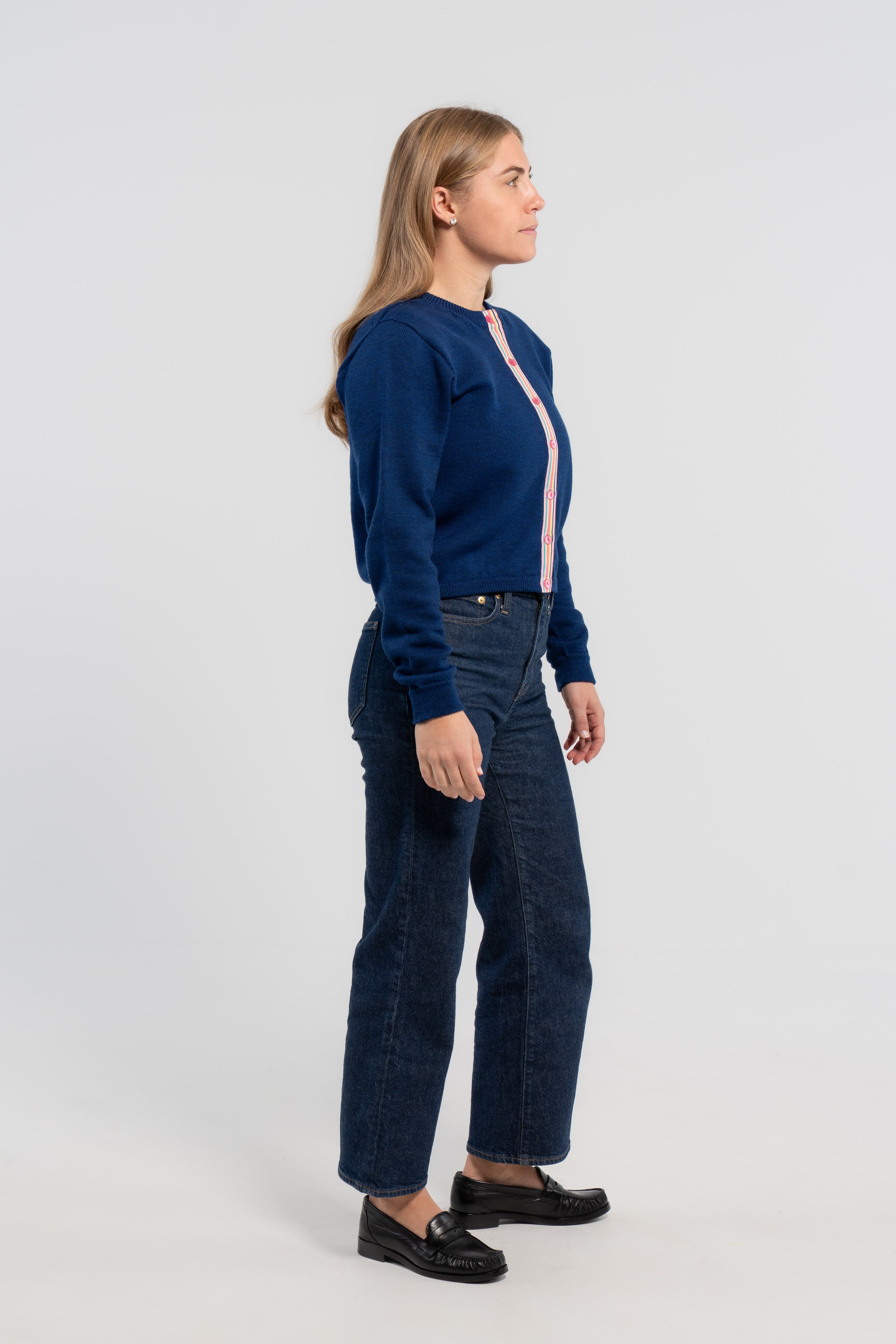 Sideview of woman wearing cobalt blue crop cardigan with pink buttons and rainbow striped grosgrain ribbon trim, jeans, loafers on plain background.