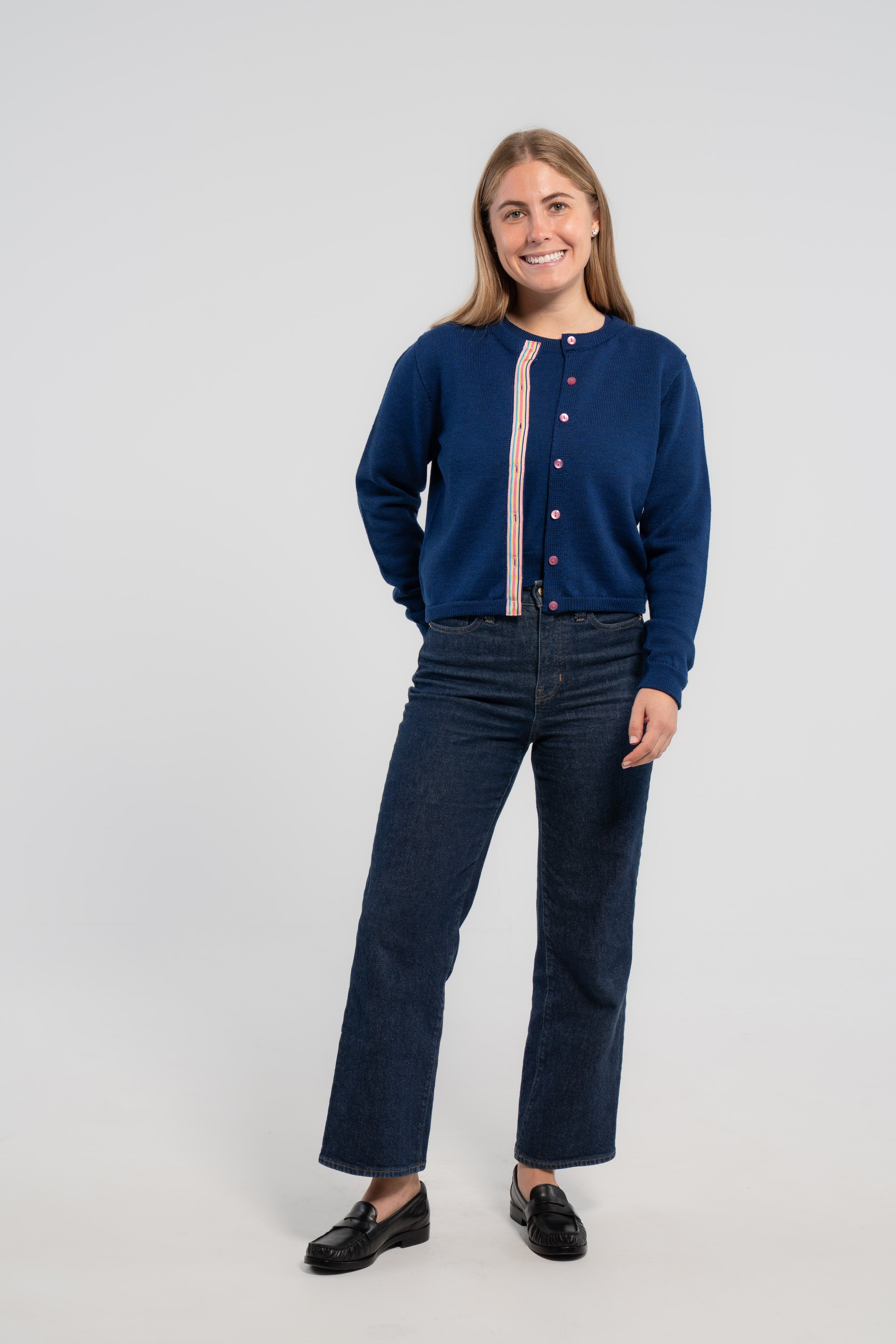 Woman wearing unbuttoned cobalt blue crop cardigan with pink buttons and rainbow striped grosgrain ribbon trim over matching cobalt blue sweater shell, jeans, loafers on plain background.