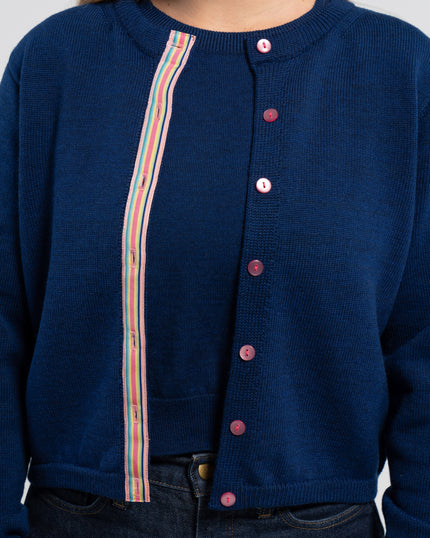 Close up of woman wearing unbuttoned cobalt blue crop cardigan with pink pearlized buttons and rainbow striped grosgrain ribbon over matching cobalt blue sweater shell and jeans.