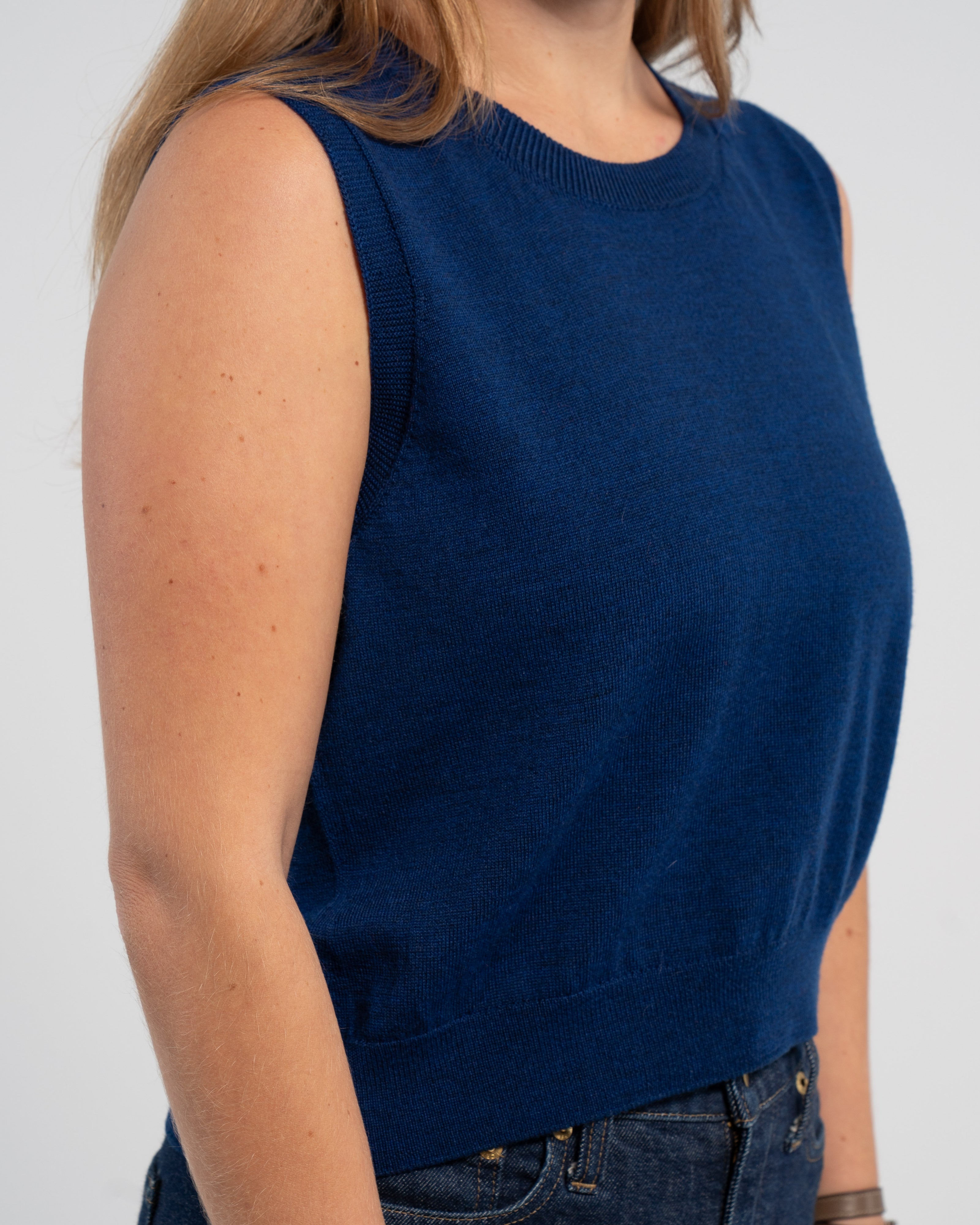 Closeup of woman wearing cobalt blue sleeveless sweater and jeans on plain background.