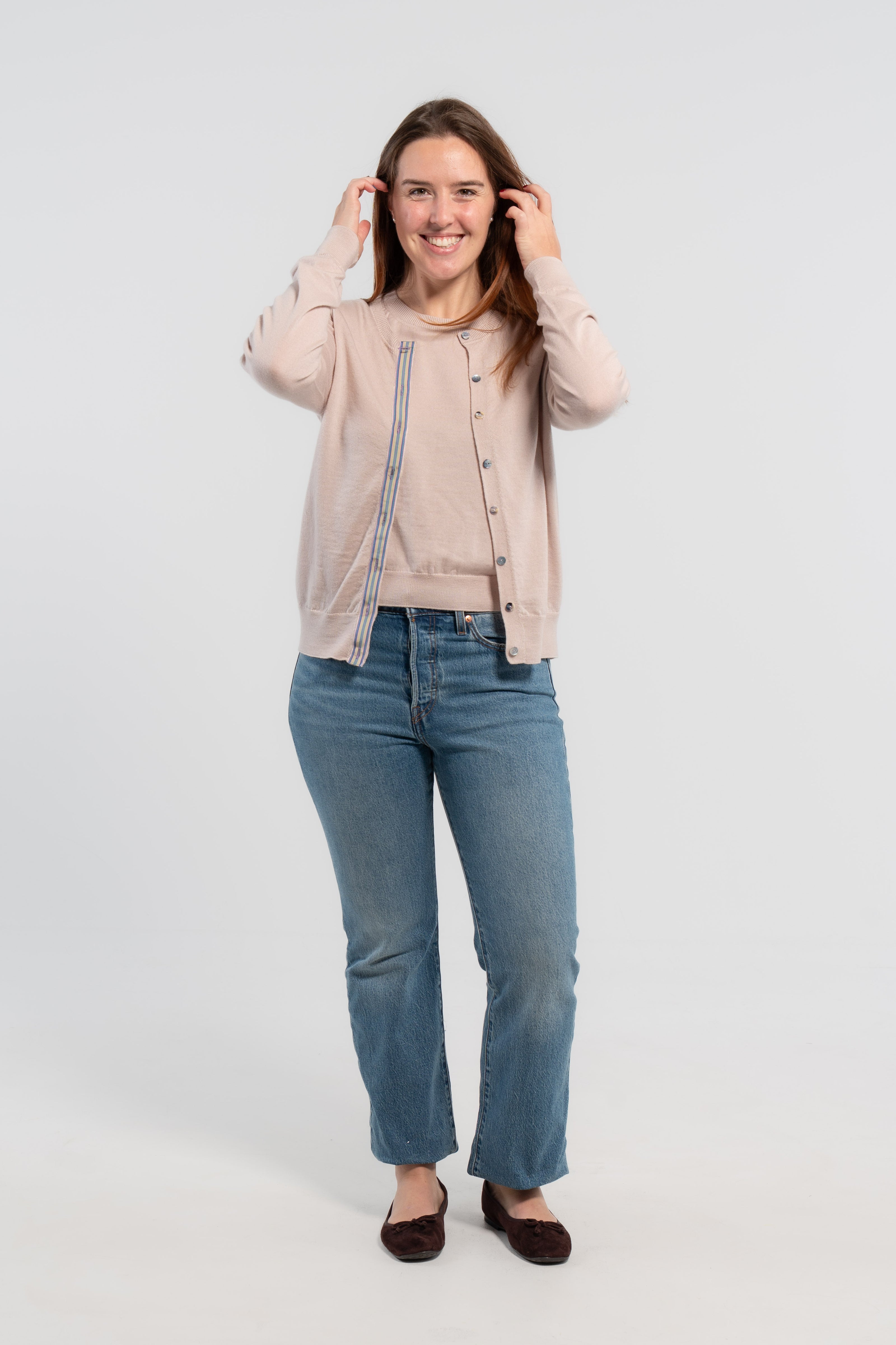 Woman wearing unbuttoned beige cardigan with iridescent shell buttons and tan, purple, lavender, and teal striped grosgrain ribbon trim over matching beige sweater shell, jeans, ballet flats on plain background.