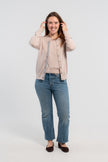 Woman wearing unbuttoned beige cardigan with iridescent shell buttons and tan, purple, lavender, and teal striped grosgrain ribbon trim over matching beige sweater shell, jeans, ballet flats on plain background.
