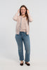 Woman wearing unbuttoned beige cardigan with iridescent shell buttons and tan, purple, lavender, and teal striped grosgrain ribbon trim over matching beige sweater shell, jeans, ballet flats on plain background.