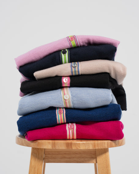Stack of colorful Noyac Knits merion wool ribbon cardigans on a wooden stool against a light gray background