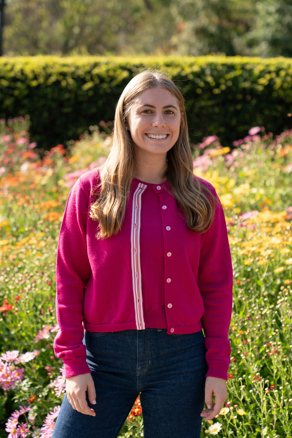 Woman wearing raspberry pink crop cardigan with pink buttons and pink, white and green striped ribbon trim and jeans with background of flowers and green hedge.