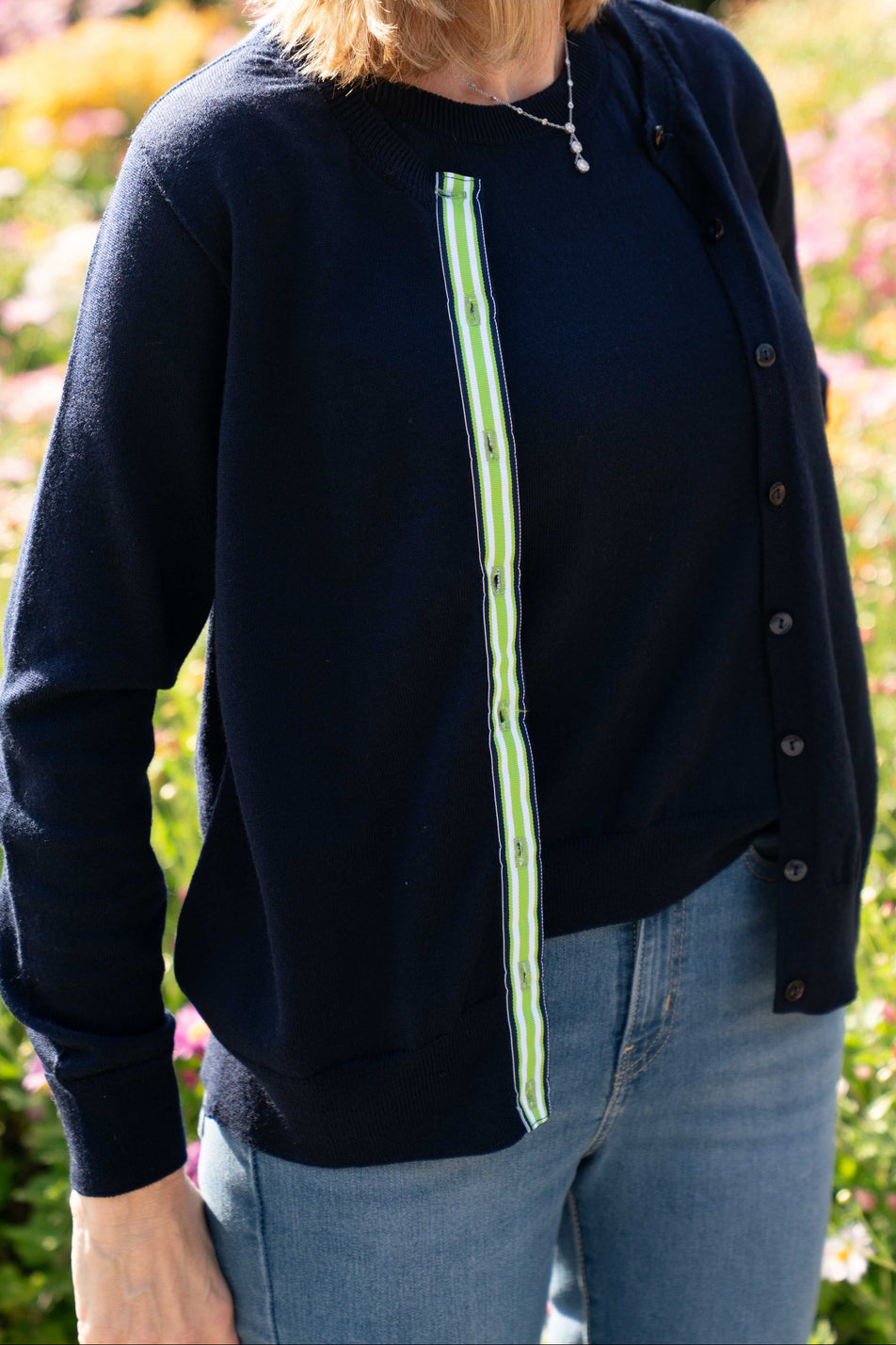 Woman wearing navy cardigan twin set with jeans and blurred flowers in the background.  Cardigan has navy, white and green striped ribbon trim.