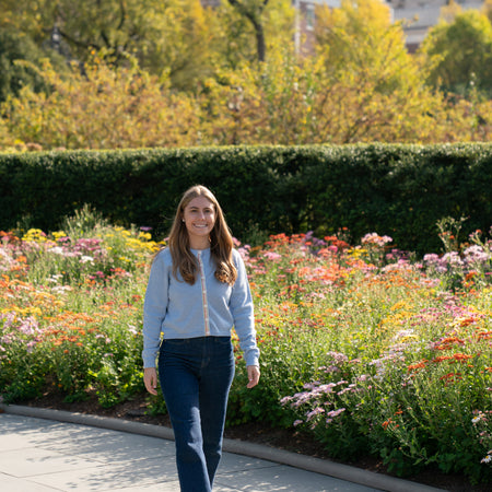 Woman wearing light blue crop cardigan with striped ribbon trim and jeans walking on a sidewalk with a garden and trees in the background.