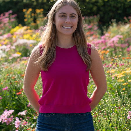 Woman in a raspberry pink sleeveless sweater and blue jeans standing in a garden with flowers and greenery.
