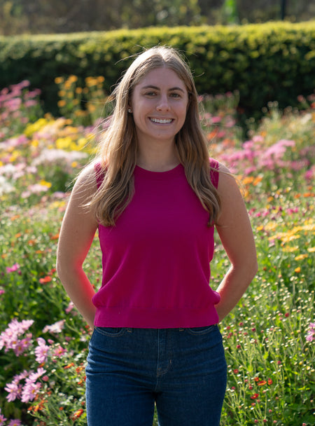 Woman in a raspberry pink sleeveless sweater and blue jeans standing in a garden with flowers and greenery.