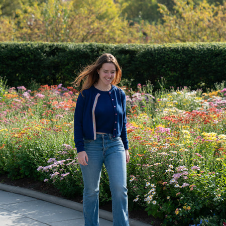 Woman walking on sidewalk in garden with green hedge and trees in background wearing cobalt blue crop sweater with pink buttons and rainbow striped ribbon trim, matching crop sleeveless sweater, jeans, ballet flats.