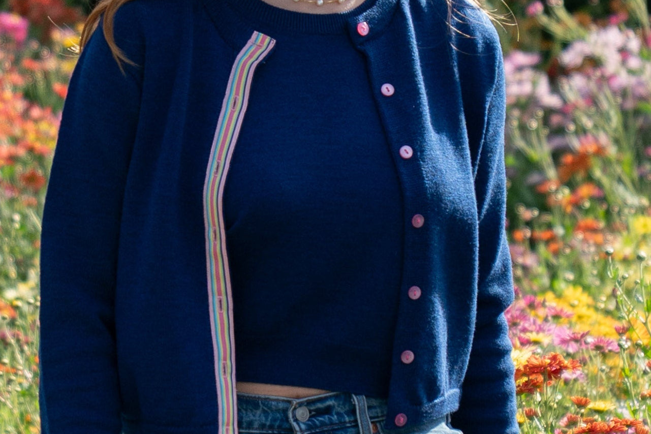 Woman walking on sidewalk in garden with green hedge and trees in background wearing cobalt blue crop sweater with pink buttons and rainbow striped ribbon trim, matching crop sleeveless sweater, jeans, ballet flats.