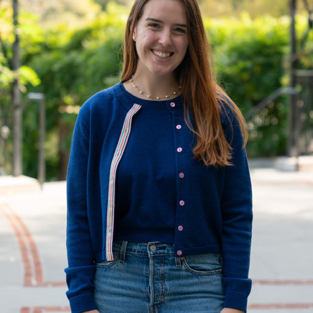 Woman on sidewalk in garden wearing cobalt blue crop cardigan with pink buttons and rainbow stripe ribbon trim over matching cobalt blue sleeveless sweater and jeans.