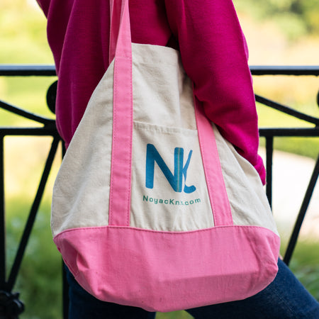 Person holding pink and white boat tote with Noyac Knits NK logo and wearing raspberry pink sweater and jeans in front of wrought iron fence with blurred greenery in backgroundl