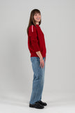 Side view of woman wearing red sweater with ribbon trim on shoulder, jeans, loafers on plain background.