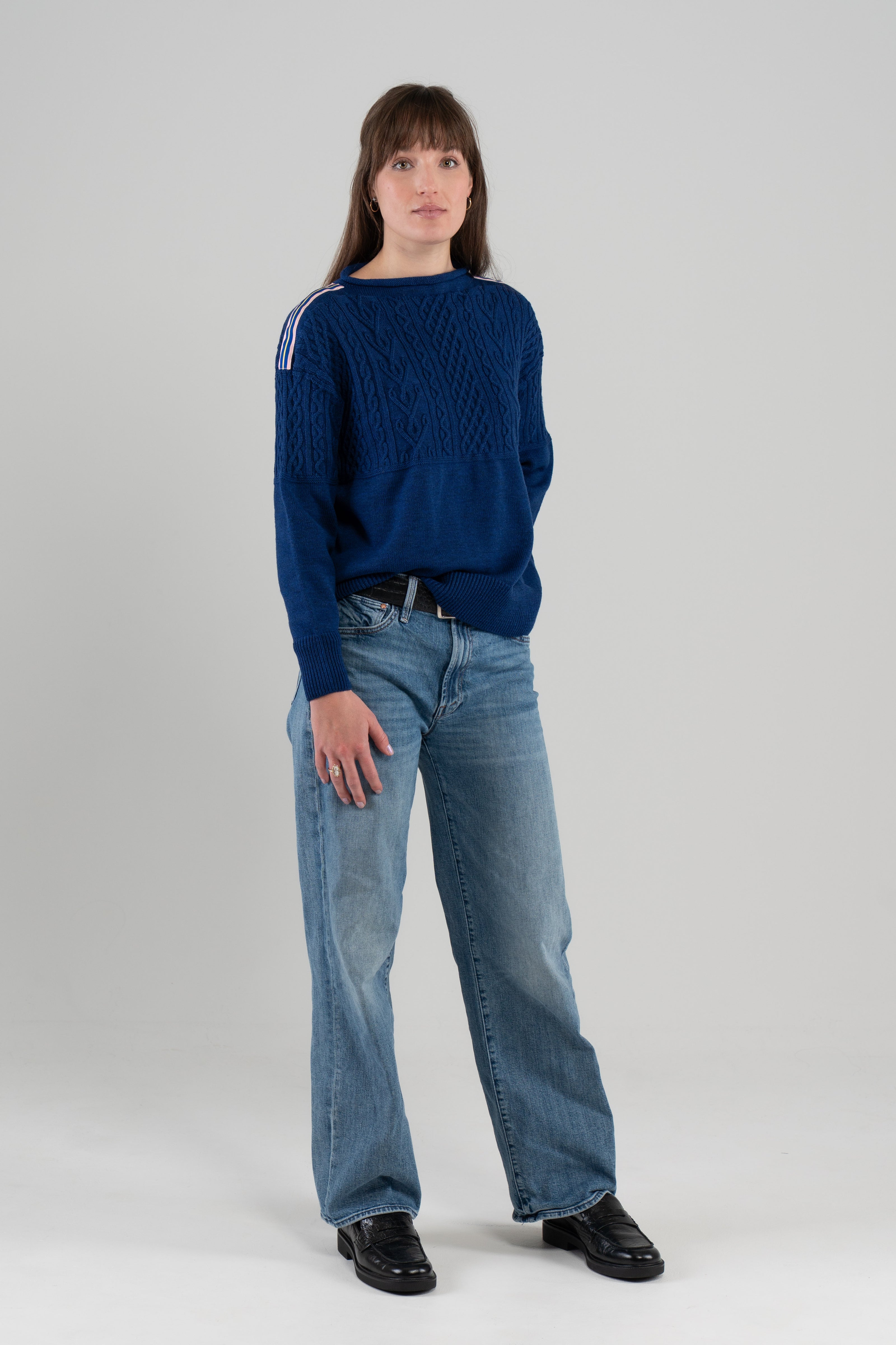 Woman wearing cobalt blue sweater with ribbon trim on shoulder, jeans, loafers and plain background.