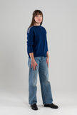 Woman wearing cobalt sweater with ribbon trim on shoulder, jeans, loafers on plain background.