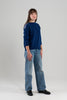 Woman wearing cobalt sweater with ribbon trim on shoulder, jeans, loafers on plain background.