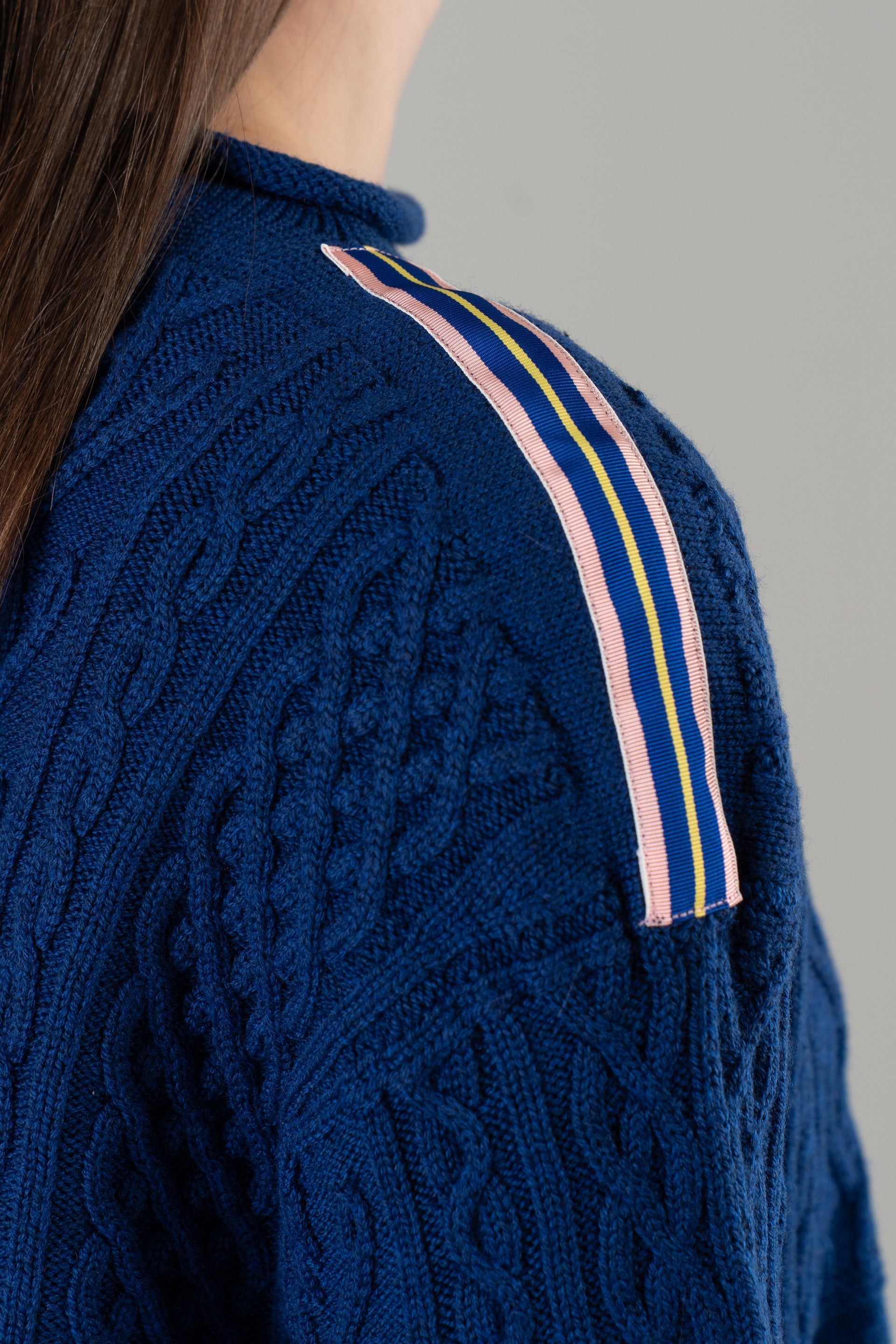 Close-up of shoulder of cobalt blue sweater with ribbon trim with pink, cobalt blue and yellow stripes on plain background.
