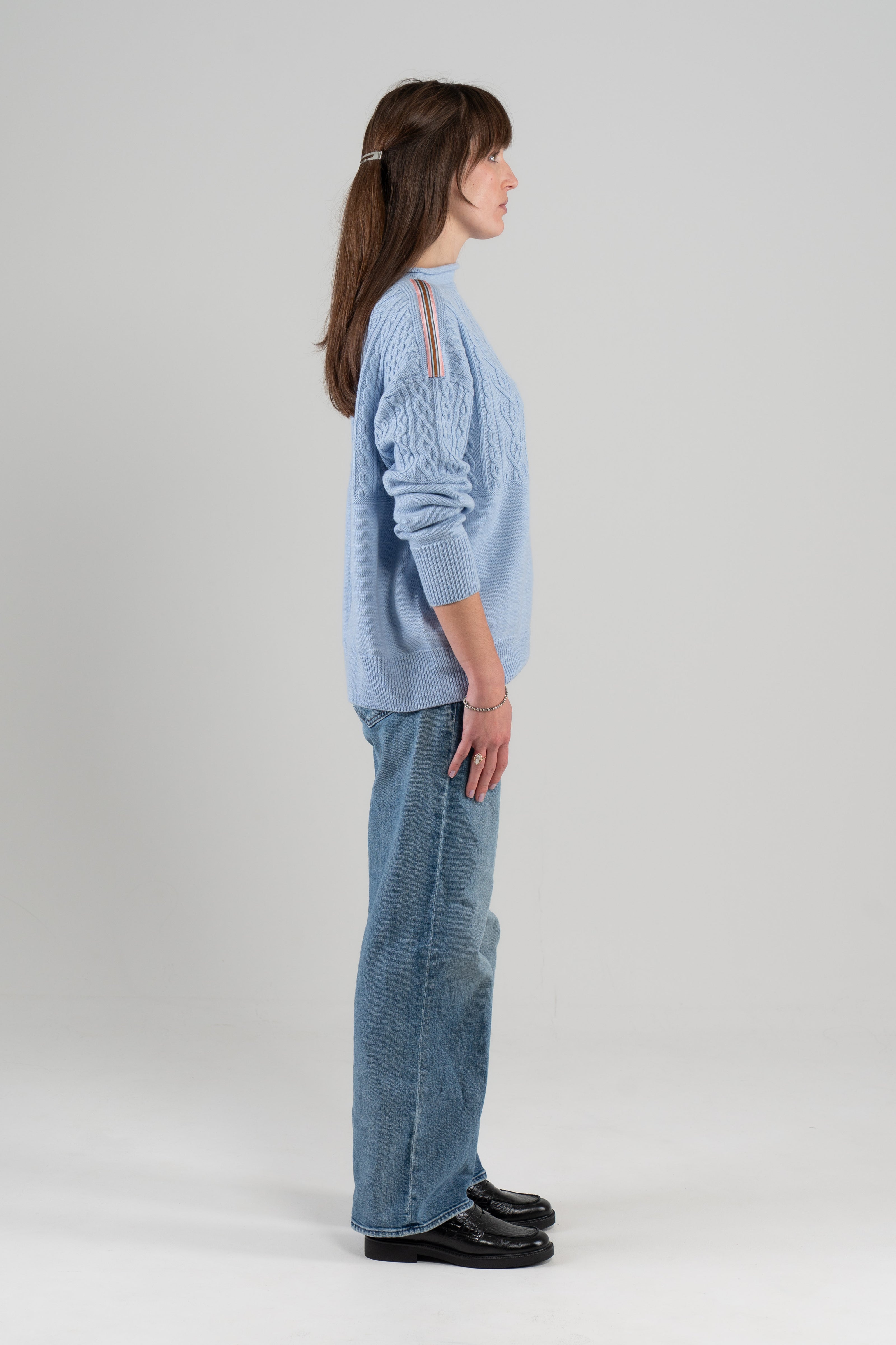 Side view of woman wearing a light blue sweater with ribbon detail on shoulder, jeans and loafers standing against a white background