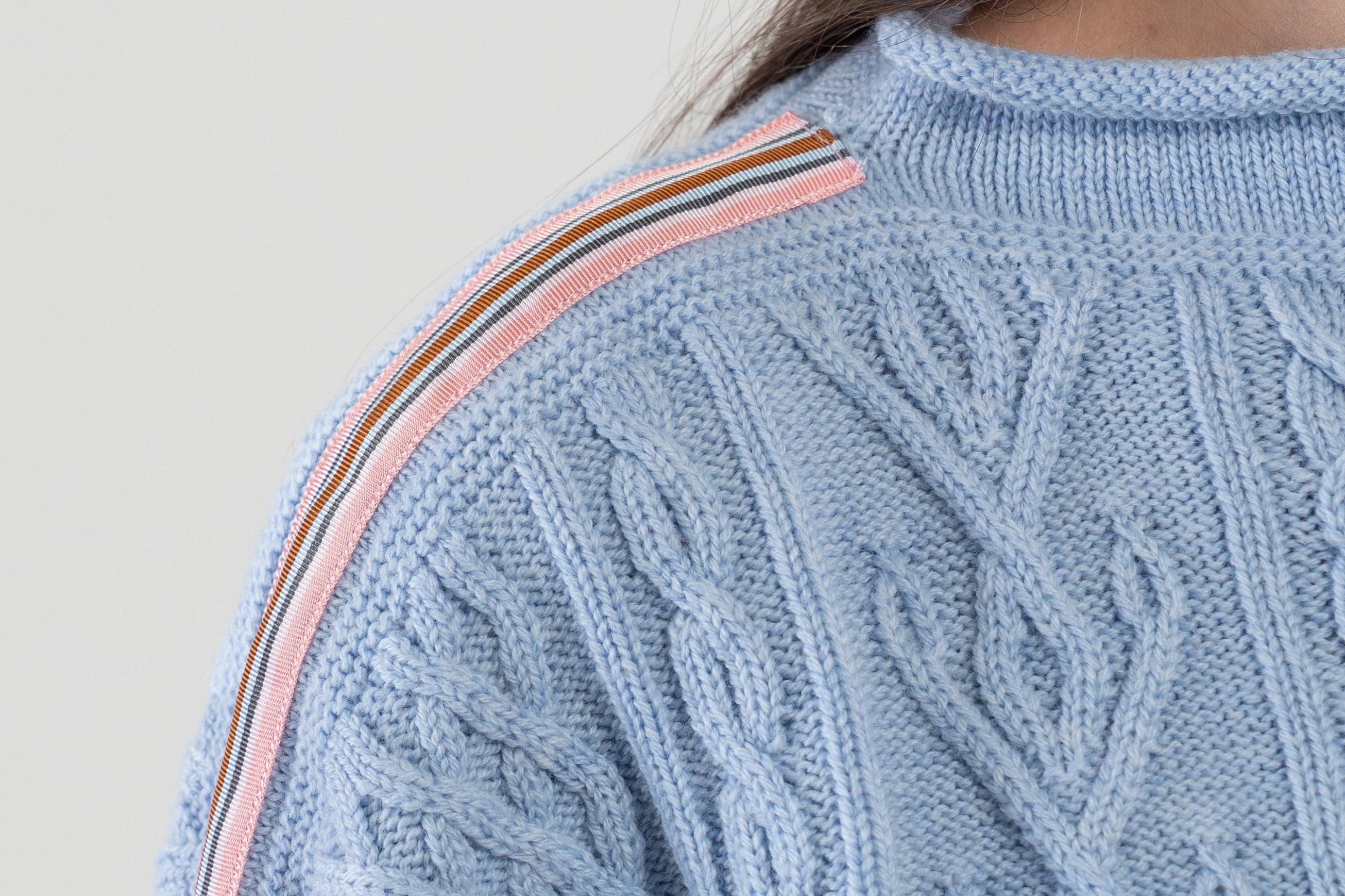 Light blue cable knit sweater with pink, navy, light blue, and brown striped ribbon trim on a plain background