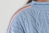 Light blue cable knit sweater with pink, navy, light blue, and brown striped ribbon trim on a plain background