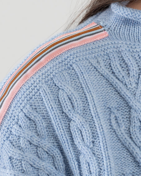 Light blue cable knit sweater with pink, navy, light blue, and brown striped ribbon trim on a plain background