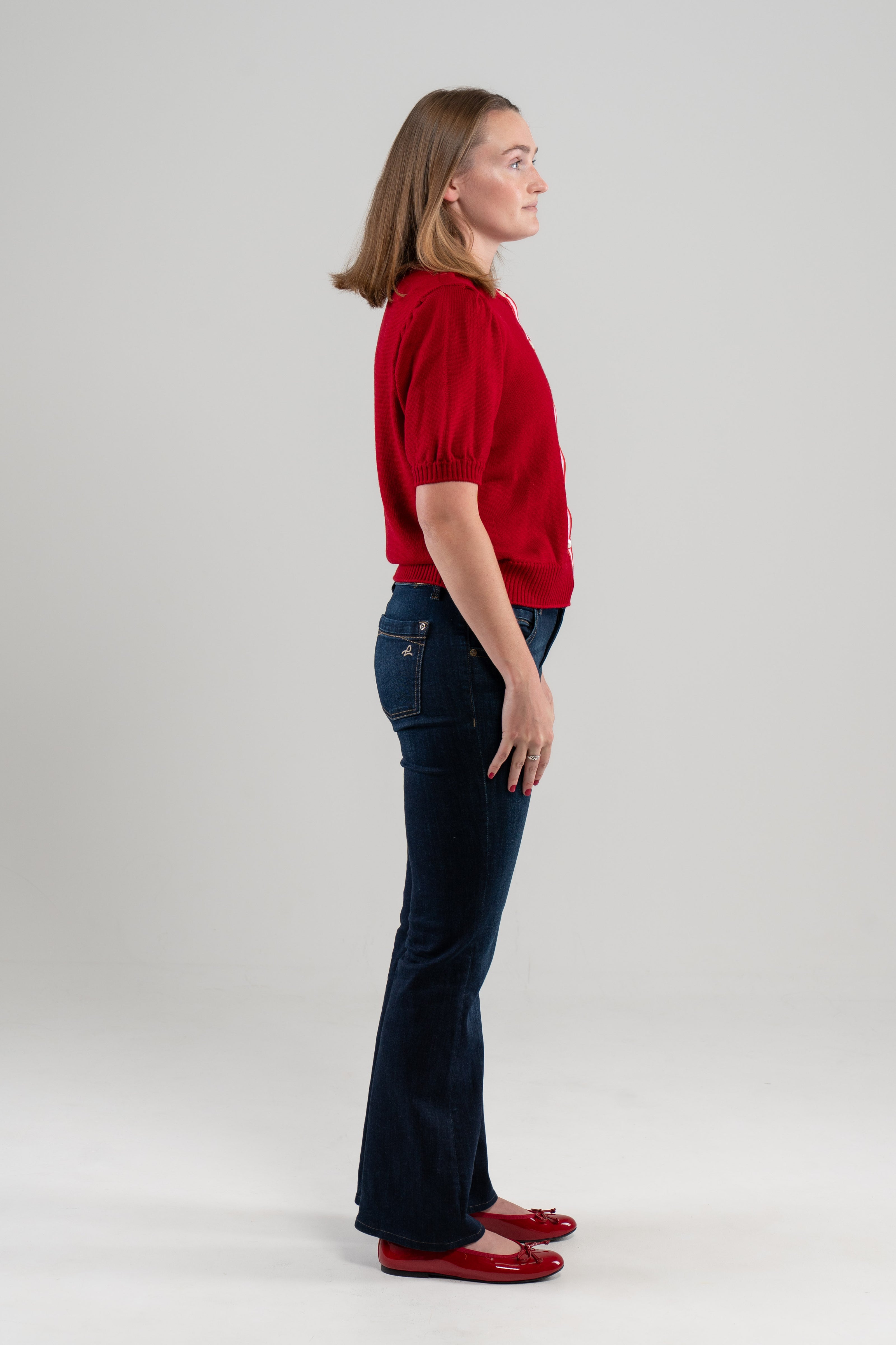 Side view of woman wearing red puff-sleeve cardigan, jeans, red shoes on plain background.