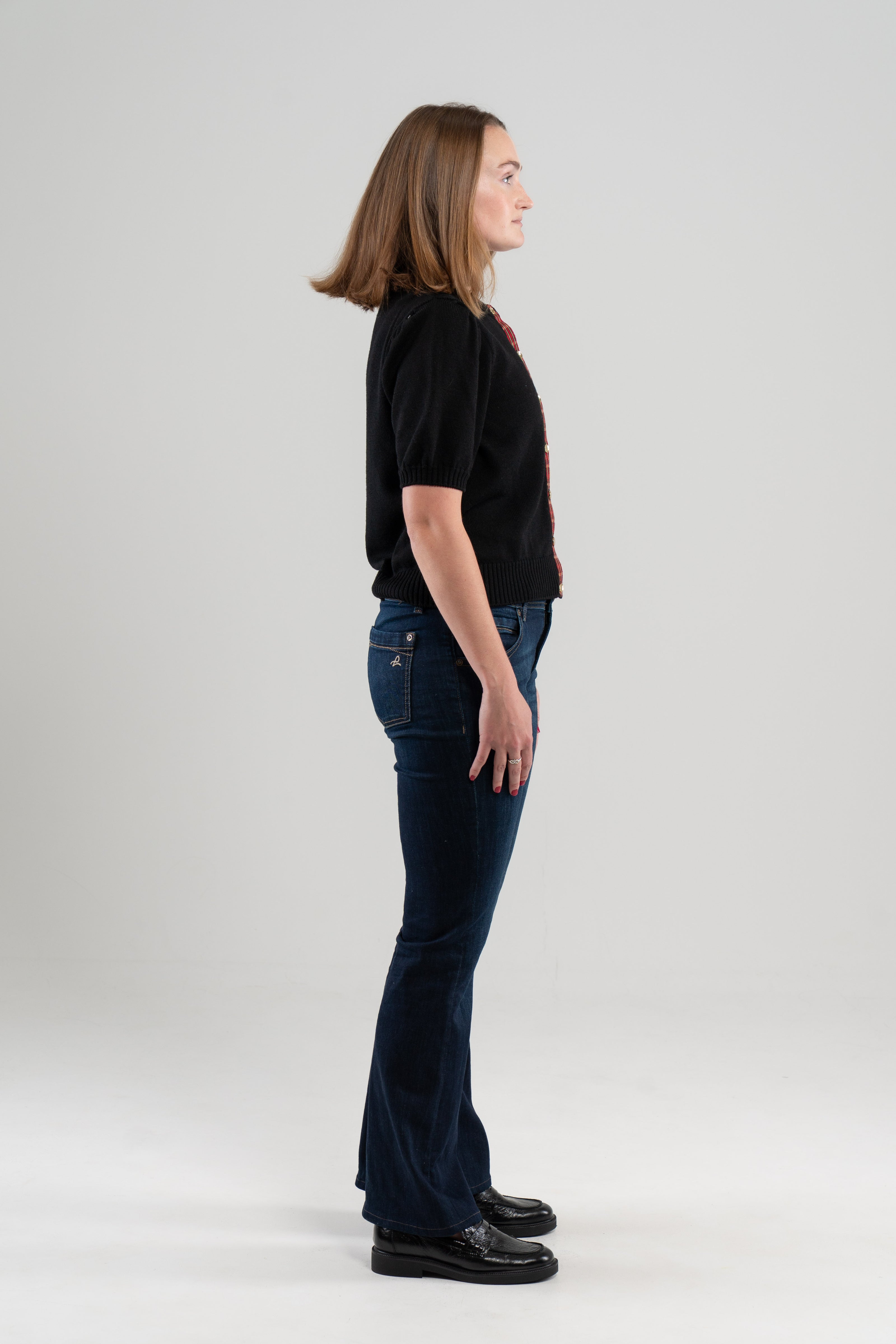 Side view of woman wearing black puff-sleeve cardigan, jeans, loafers on plain background.