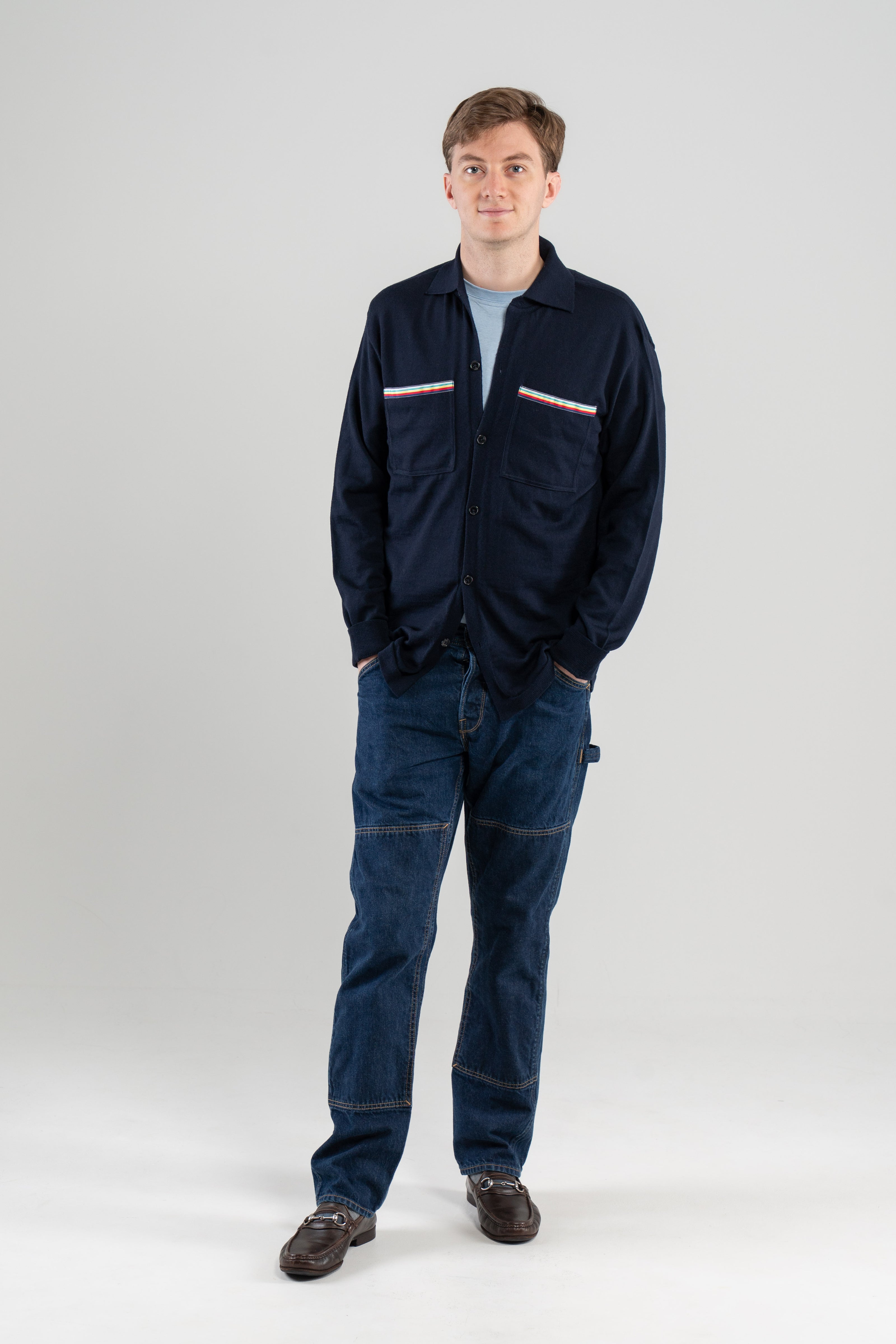 Man wearing navy overshirt with striped ribbon trimmed chest pockets over light blue tee-shirt, jeans, loafers on plain background.