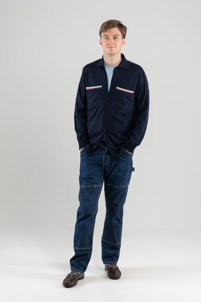 Man wearing navy overshirt with striped ribbon trimmed chest pockets over light blue tee-shirt, jeans, loafers on plain background.