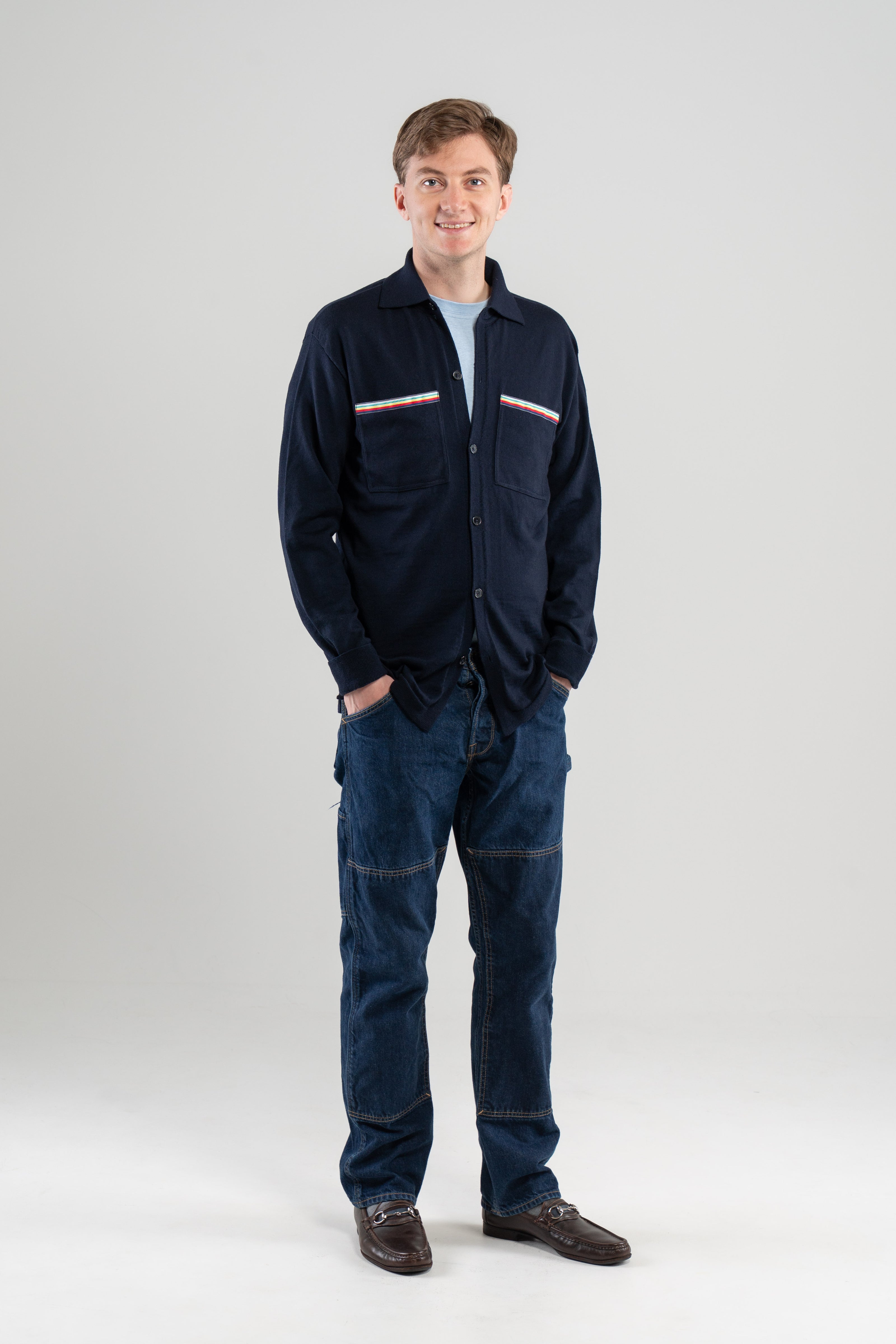 Partial side view of man wearing navy overshirt with striped ribbon-trimmed chest pockets over light blue tee-shirt, jeans, loafers on plain background.