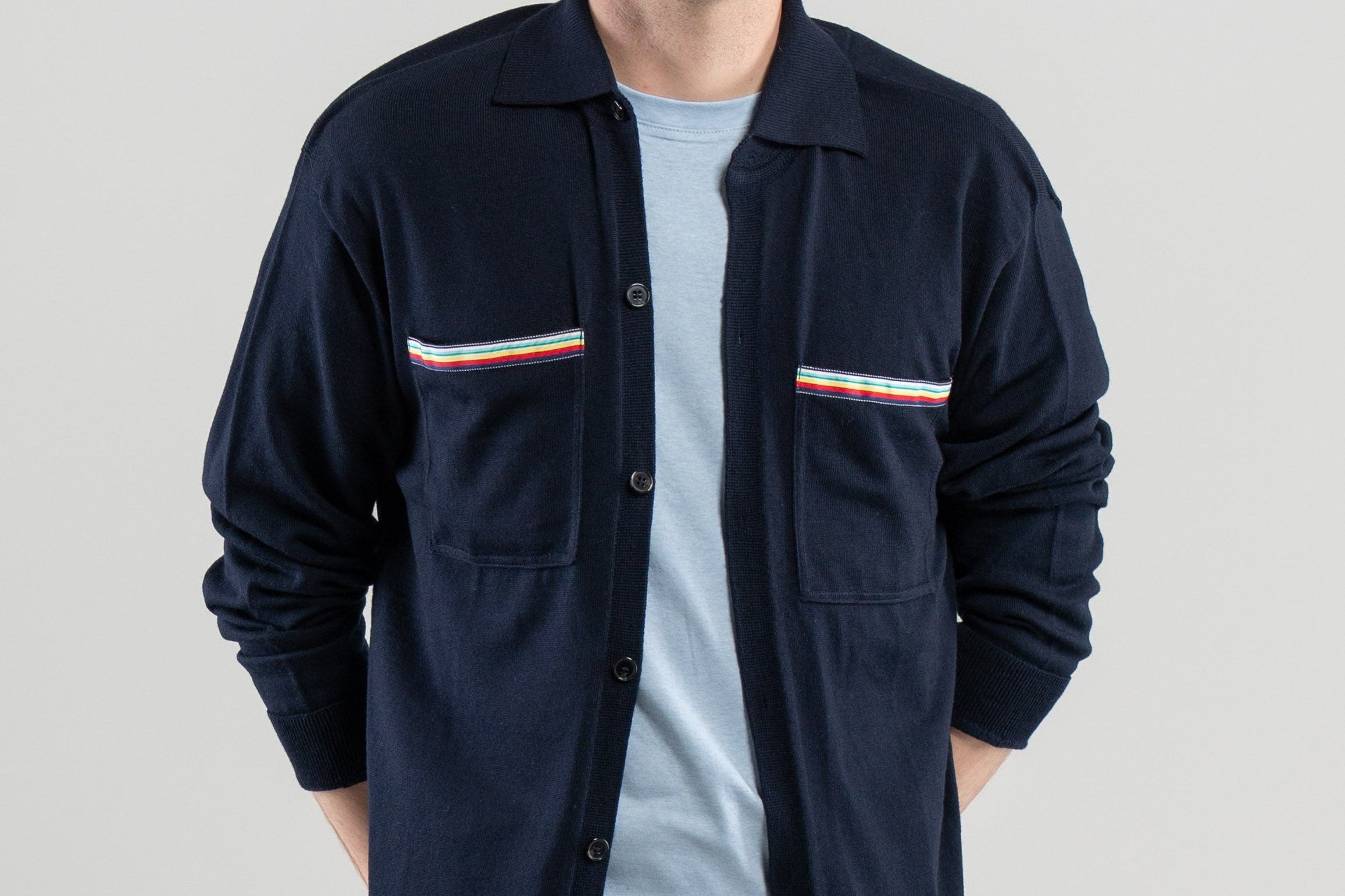 Man wearing unbuttoned navy overshirt with striped ribbon trimmed chest pockets, light blue tee-shirt, jeans, loafers on plain background.