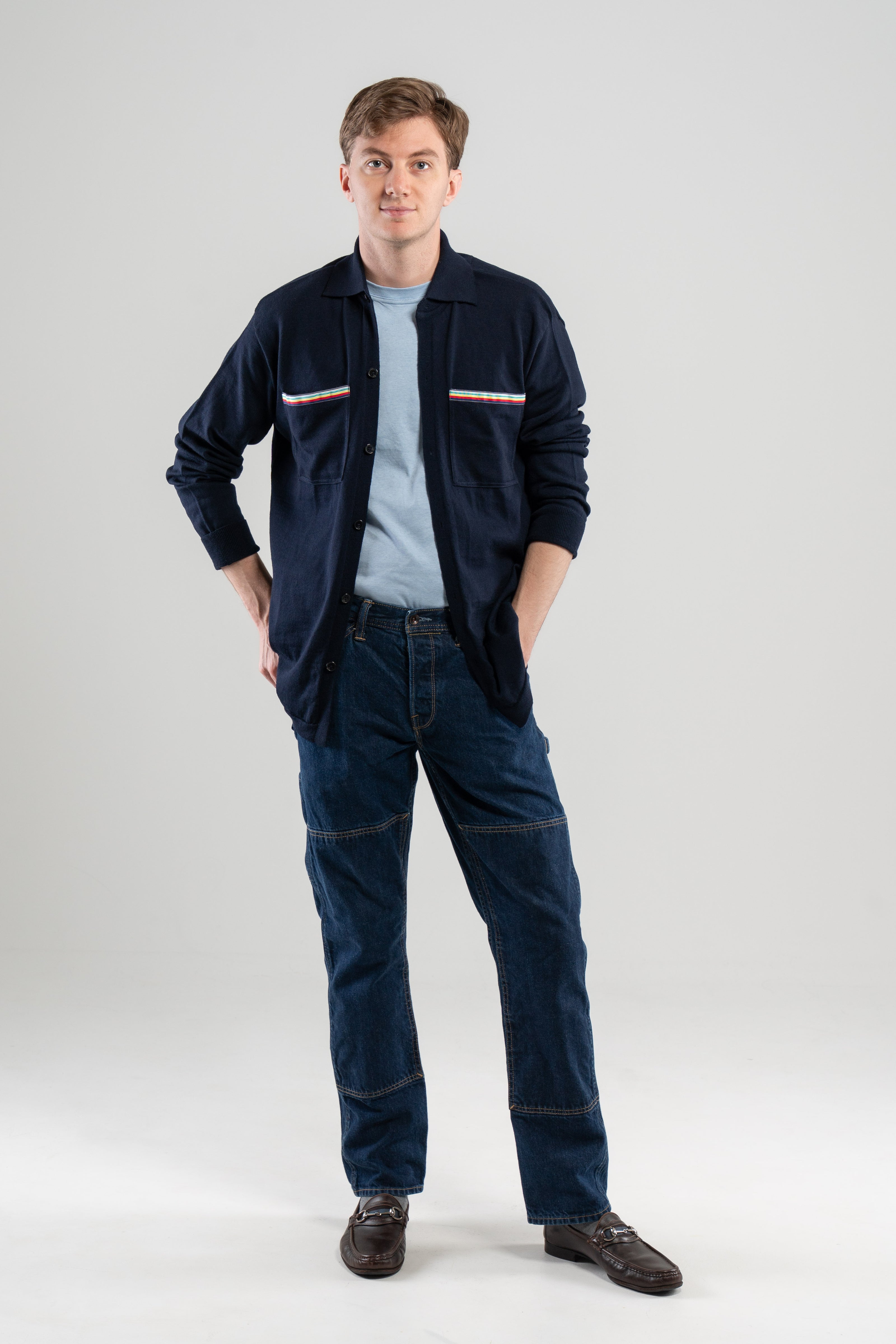 Man wearing navy overshirt with striped ribbon trimmed chest pockets, light blue tee-shirt, jeans, loafers on plain background.