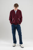 Man looking down wearing burgundy zip-up sweater with striped ribbon-trimmed pockets, jeans, sneakers on plain background.