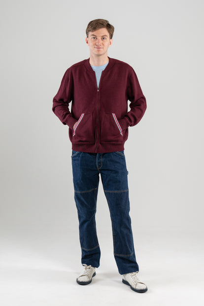 Man wearing burgundy sweater with striped ribbon-trimmed pockets and hand in pockets.  Sweater is worn over light blue tee-shirt.  Man is wearing jeans and sneakers.  Plain background.