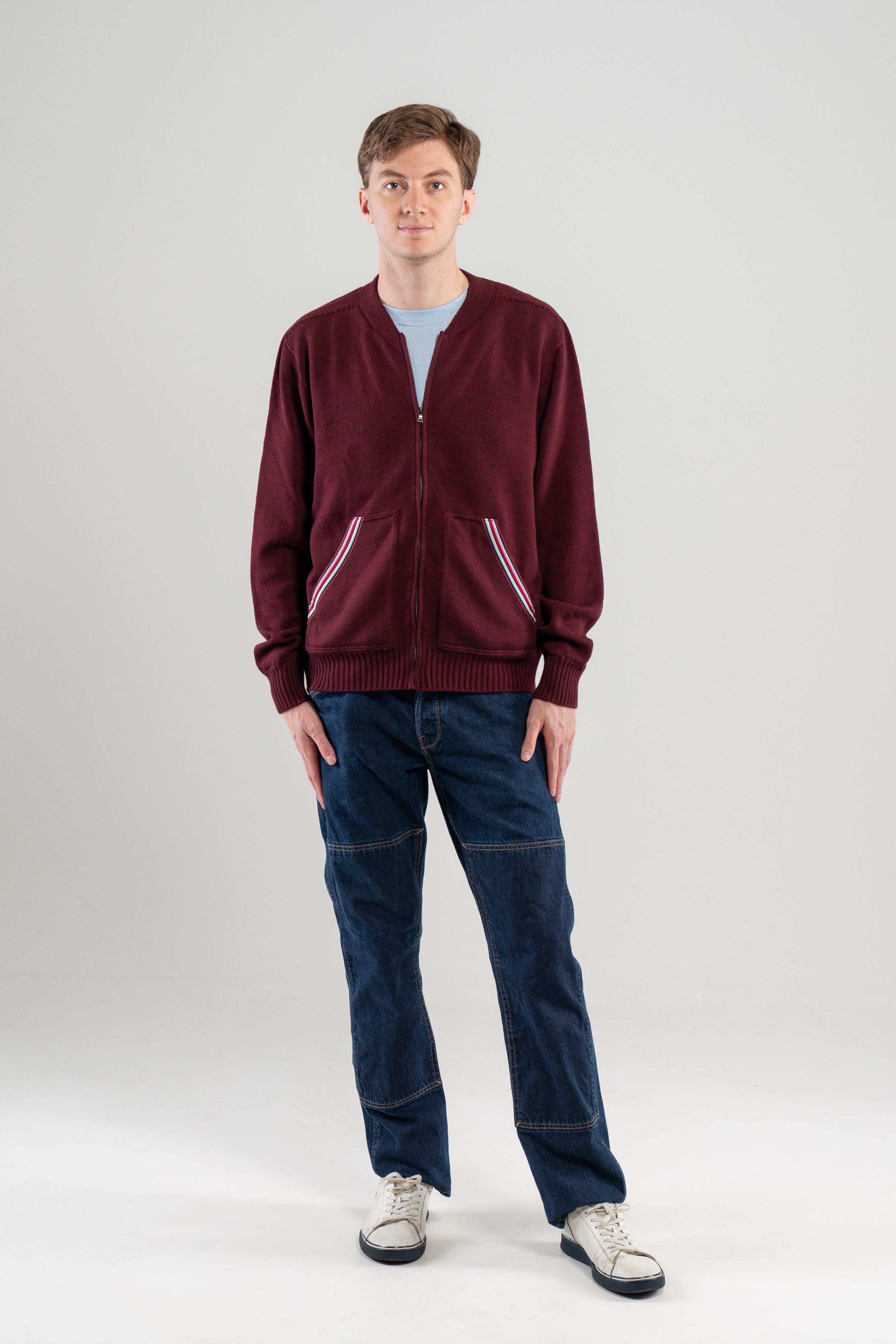 Man wearing burgundy zip-up sweater with ribbon-trimmed pockets over light blue tee-shirt, jeans, sneakers on plain background.