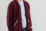 Side view of man wearing unzipped burgundy cardigan with striped ribbon trimmed pockets over light blue tee-shirt, jeans, sneakers and plain background.