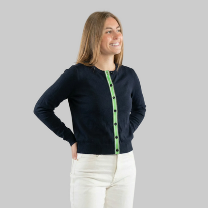 Woman wearing a navy cardigan with green and white striped ribbon trim and navy styled wirh white jeans on a white background