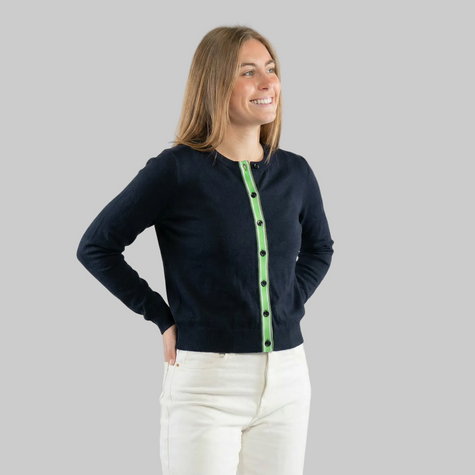 Woman wearing a navy cardigan with green and white striped ribbon trim and navy styled wirh white jeans on a white background