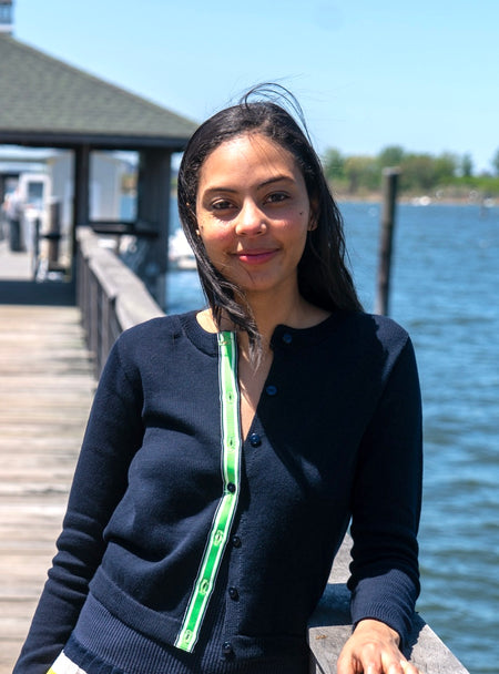 Woman standing on a dock by the bay wearing Noyac Knits navy Julie crop cotton cardigan trimmed with signature ribbon in green, white and navy stripes -- knit in premium Egyptian cotton.