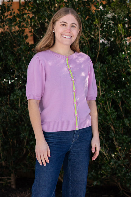 Woman wearing Noyac Knits lilac pink short puff-sleeve cardigan with pink and green ribbon trim, in premium, lightweight merino, styled with jeans against green leafy background