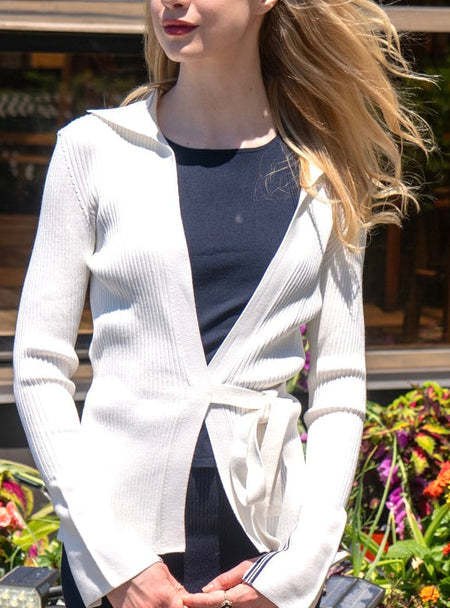 Woman in Noyac Knits white wrap cardigan with ribbon-trimmed sleeves styled with Noyac Knits ribbed knit stretch midi skirt in navy and navy scoop neck tank, all in ecoviscose.  Woman is  standing in front of a garden with flowers and plants.