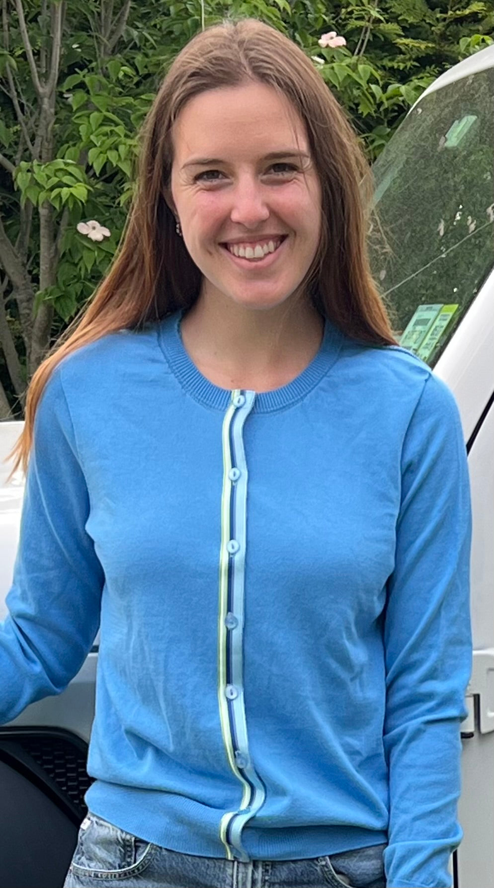 Woman wearing Noyac Knits Allison classic cardigan in sky blue premium cotton and trimmed with green and blue striped ribbon in natural setting outdoors